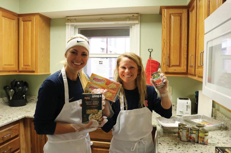 Two softball players volunteer in a kitchen.